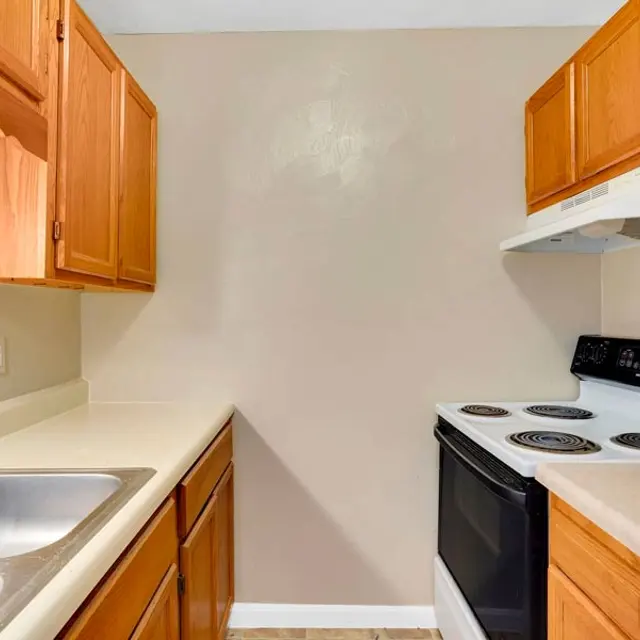 A small kitchen with wooden cabinets, a double sink, a countertop, and a black stove. The walls are painted light beige, creating a warm atmosphere.