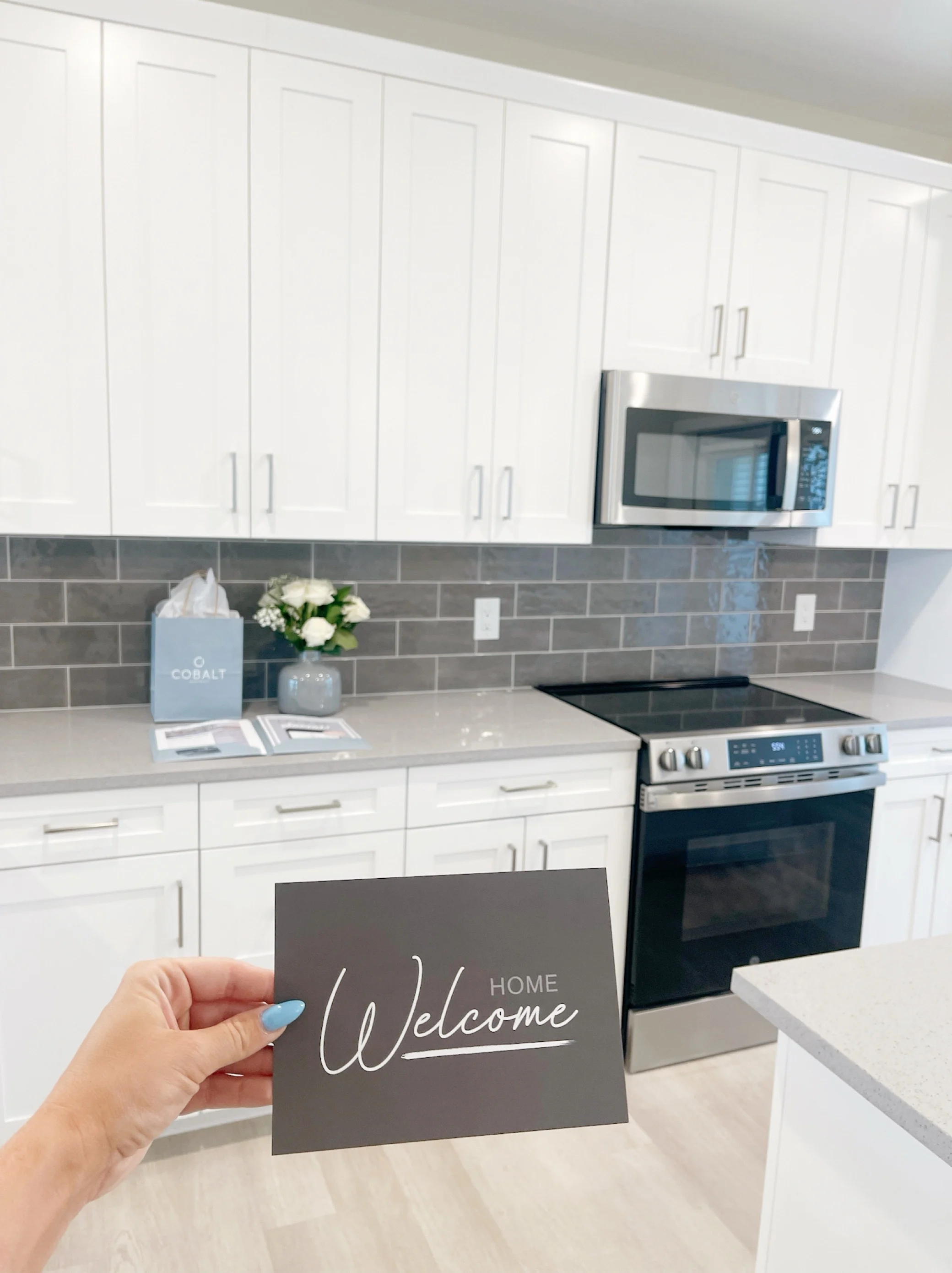 Welcome Home to a Modern Kitchen A modern kitchen with white cabinets and grey tile backsplash. A hand is holding a 'Welcome Home' card in the foreground, with flowers and a gift bag on the counter.