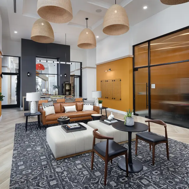A modern living area featuring a brown leather sofa, a white ottoman coffee table, and a patterned rug. The space has large windows and decorative pendant lighting, with orange and neutral walls.