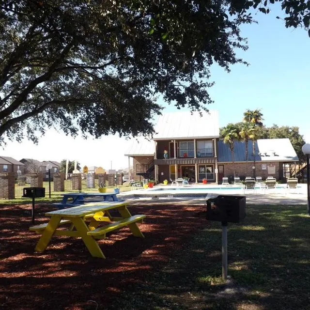 A view of a swimming pool area featuring lounge chairs, a picnic table, and barbeque grills surrounded by greenery and residential buildings in the background.