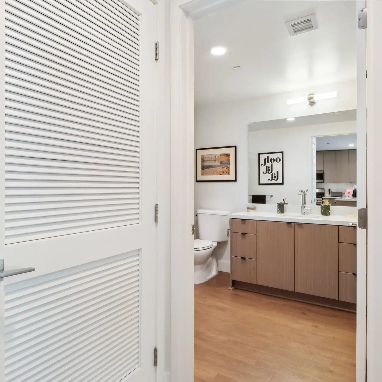 A modern bathroom interior seen through an open door, featuring a large mirror, sink, and wooden cabinetry with a graphic art piece on the wall.