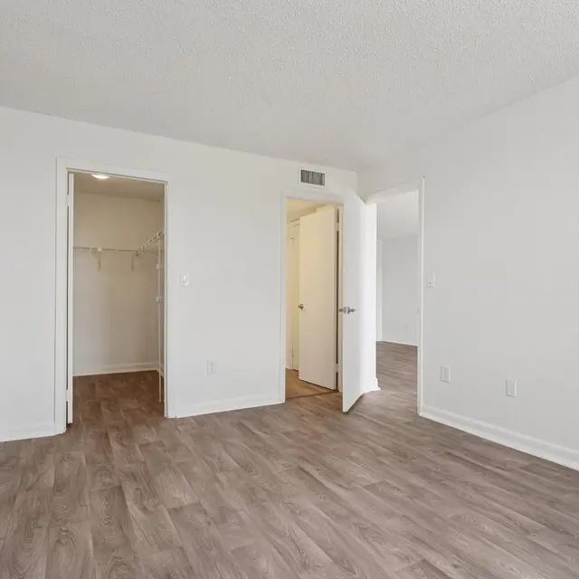 Bright and Spacious Empty Room A spacious, empty room with light-colored walls and a wooden floor. Two doorways are visible; one leads to a closet and the other to an adjacent room. Natural light fills the space, creating a bright atmosphere.