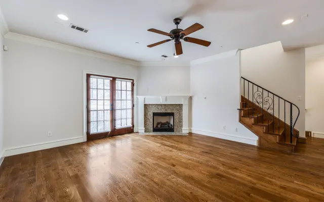 Contemporary Living Room Interior A spacious living room featuring a fireplace, wooden flooring, a ceiling fan, and a staircase. The room has large windows with French doors, allowing natural light to fill the space.