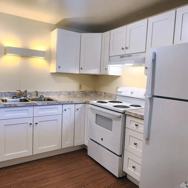 An interior view of a modern kitchen featuring white cabinetry, a granite countertop, a white stove, refrigerator, and brown wood-like flooring.