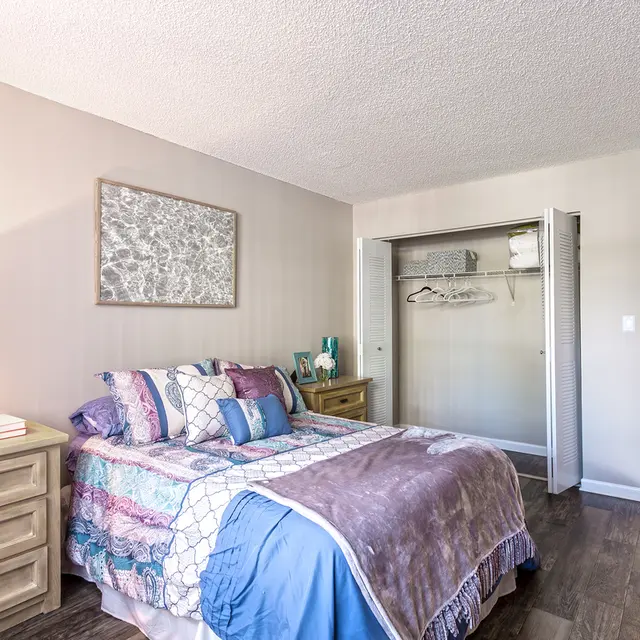 Bedroom - Biscayne Apartments A cozy bedroom featuring a double bed with a colorful patchwork blanket, a nightstand with a lamp, and an open closet with some items hanging inside.