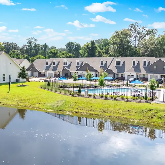Apartment Complex with Pool and Pond View of an apartment complex with a swimming pool surrounded by green lawns and trees, with a pond in the foreground under a blue sky.