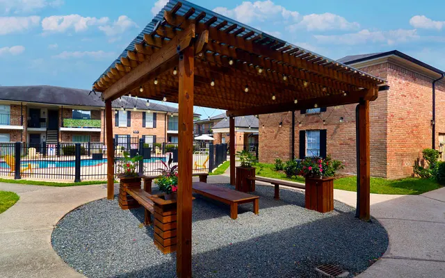 A wooden pergola surrounded by gravel and green grass, with benches and flower planters beneath it, in a residential area.
