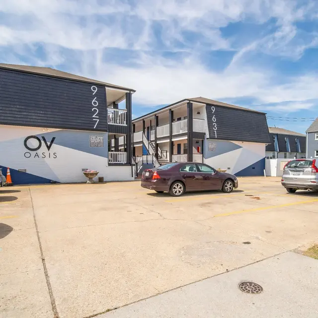 A view of the OV Oasis building, featuring two separate units, one with a blue and white facade and the other with a contrasting design. In front, there is a parking area filled with cars and a sign indicating the property name.