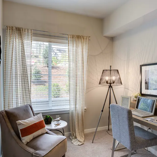 A cozy home office space featuring a small desk with a laptop and a chair, a floor lamp, and a large window with sheer curtains. Decorative items are on the desk and a comfortable armchair is present in the corner.