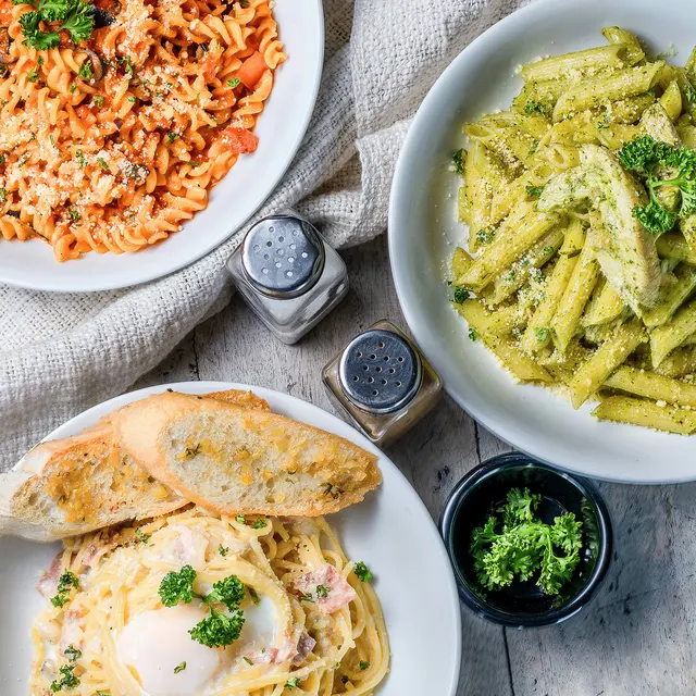 La Carafe Three plates of pasta dishes with garlic bread, featuring colorful rotini, pesto pasta, and creamy spaghetti with an egg on top.