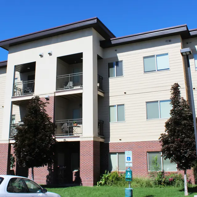 A modern three-story apartment building with a mix of large windows and balconies. The structure features a tan exterior with brick accents and is surrounded by green grass and small trees. A couple of cars are parked in front of the building.