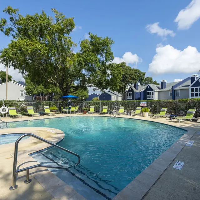 A clean swimming pool surrounded by lounge chairs, with a backdrop of residential buildings and trees under a clear blue sky.