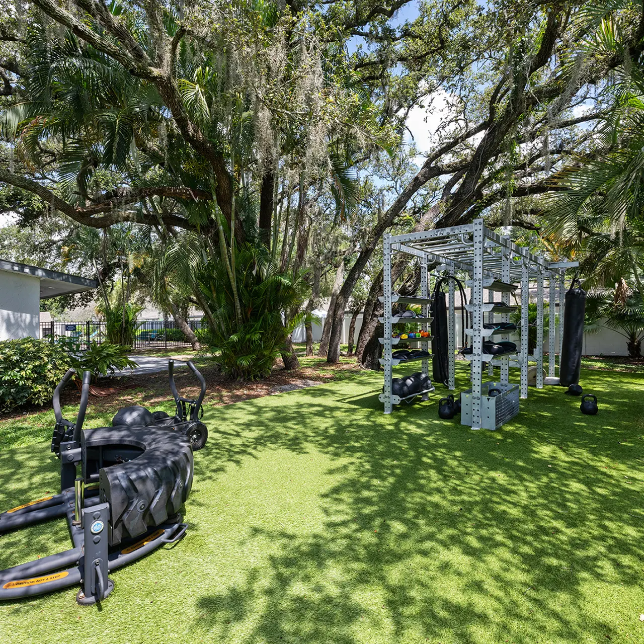 An outdoor gym setup featuring a variety of fitness equipment including a sled and a multi-station rack, surrounded by lush greenery and trees.