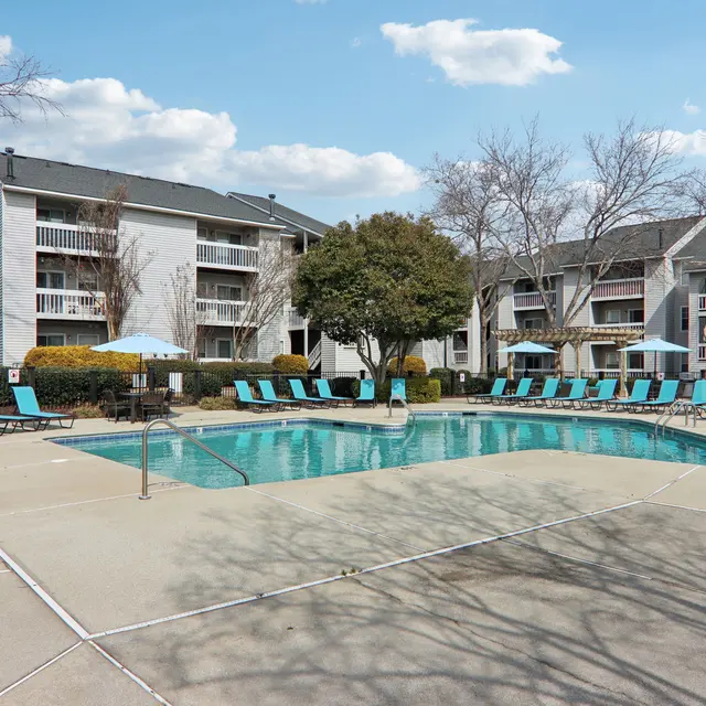 A residential apartment complex featuring a swimming pool with loungers and umbrellas. Trees are in the background, and the sky is mostly sunny with scattered clouds.