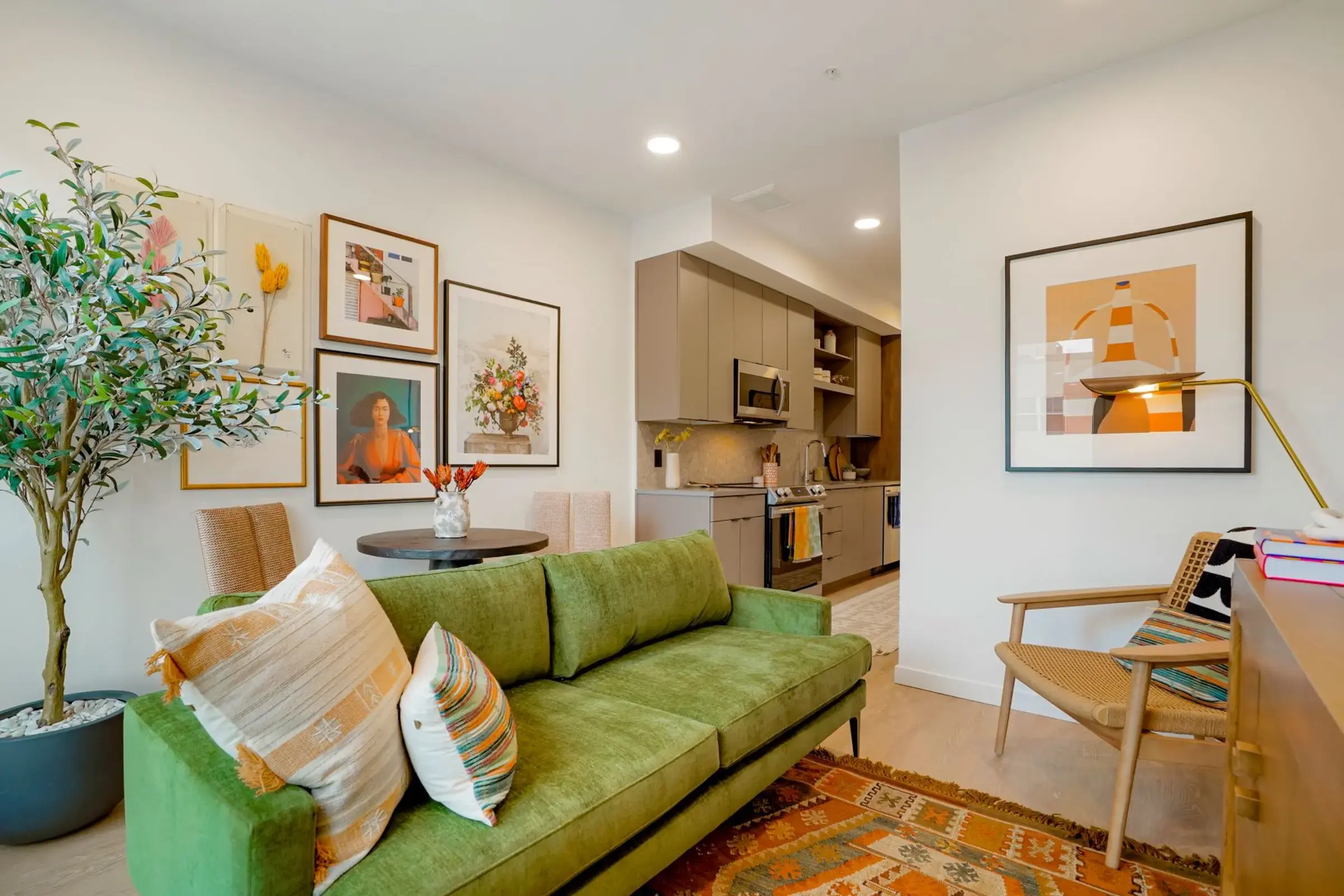 A cozy living room with a green sofa, framed artwork on the walls, a small dining table with flowers, and a view of a kitchen area.