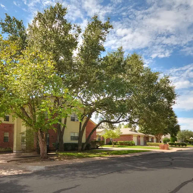 A residential street view featuring two-story houses with green lawns and large trees. The sky is partly cloudy with blue patches. The street is paved with visible cracks.