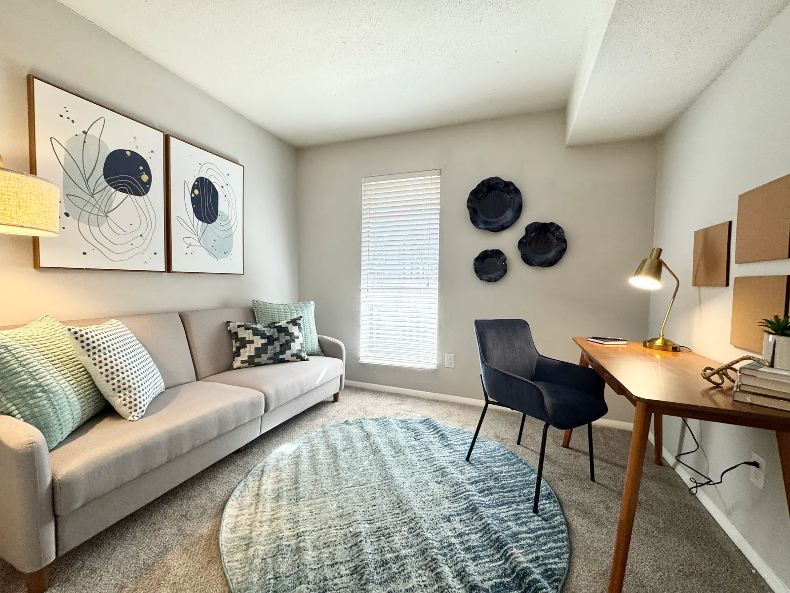 A cozy living space featuring a light gray couch adorned with various textured pillows, a round blue area rug, a large window with blinds, a wooden desk with a lamp, and wall decorations including framed art and circular wall hangings.