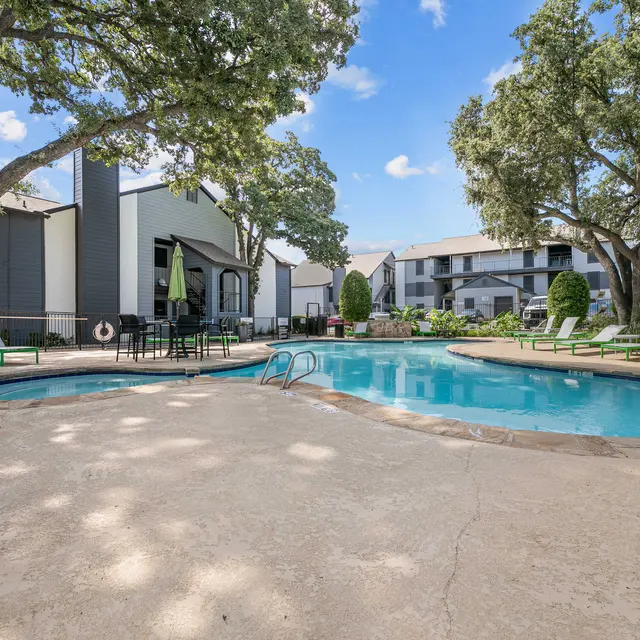 View of a swimming pool area surrounded by trees and lounge chairs, with apartment buildings in the background.