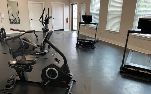 Interior of a modern gym with various workout equipment including a stationary bike, treadmill, and elliptical machine, large windows allowing natural light, and a minimalist design.