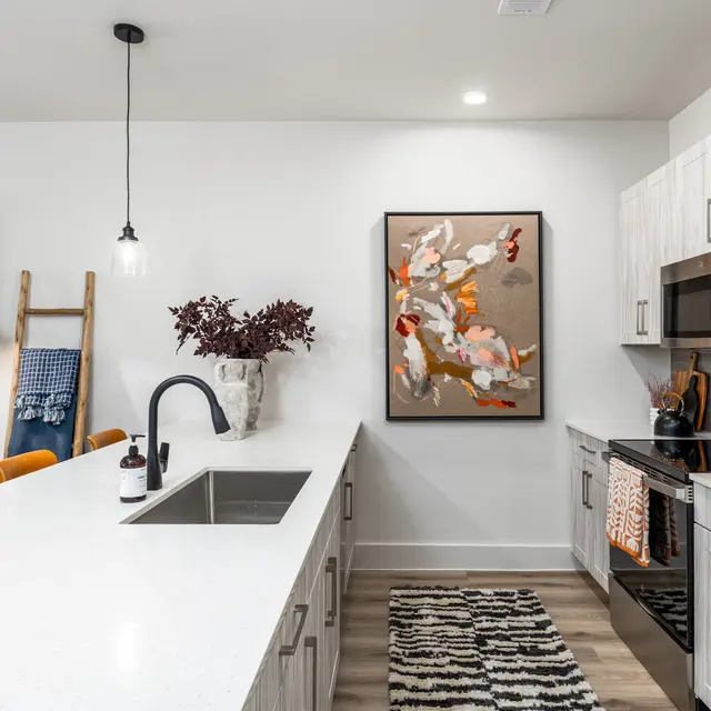 A modern kitchen featuring a large island with a white countertop, stainless steel appliances, and a decorative piece on the wall. There is a rug on the floor and stylish lighting fixtures above the island.
