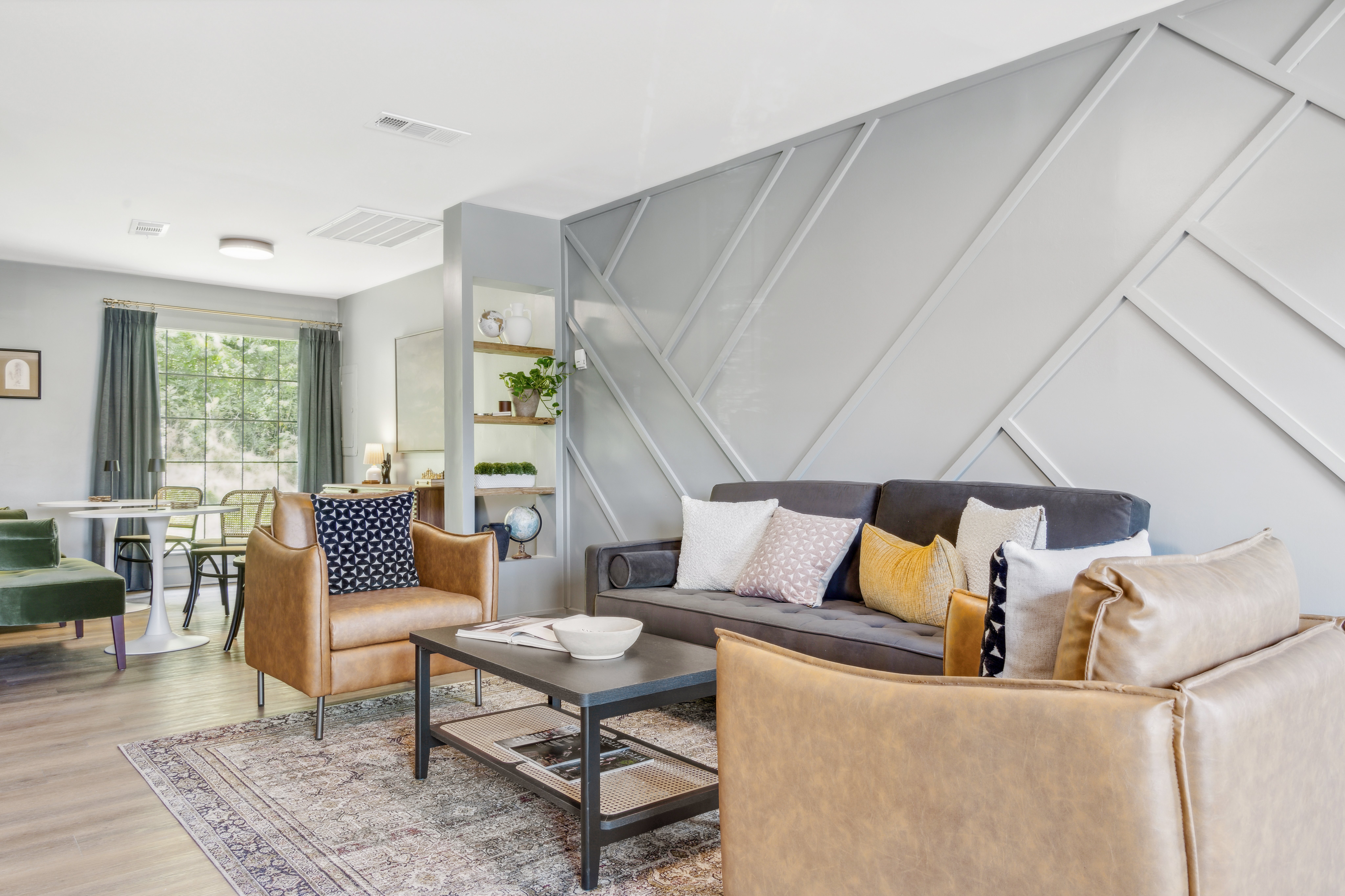 The Easton A modern living room featuring a sofa, armchairs, a coffee table, and a large window letting in natural light. Decorative pillows in various colors are arranged on the sofa, and there is a patterned rug on the floor.