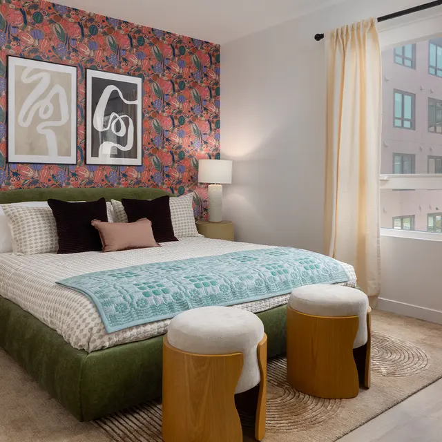 A stylish bedroom featuring a vibrant floral accent wall, a large bed adorned with decorative pillows, two bedside lamps, and a view through large windows. The decor includes a round plant pot and modern design elements.