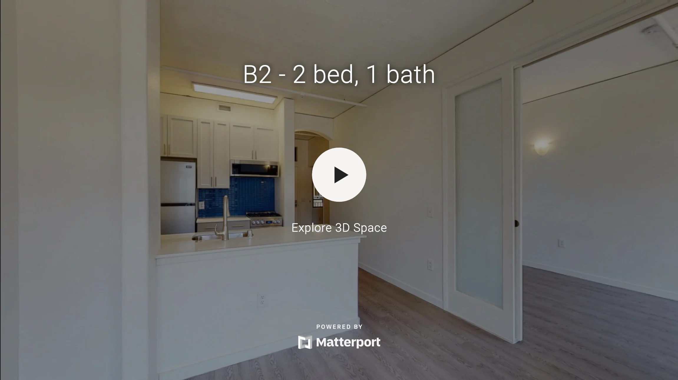 Interior view of a modern apartment showing a kitchen area and living space with light-colored walls and wood flooring.