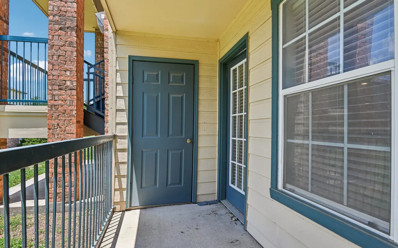 An exterior view of an apartment entrance featuring a double door and a window, with a railing and stairway visible in the background.