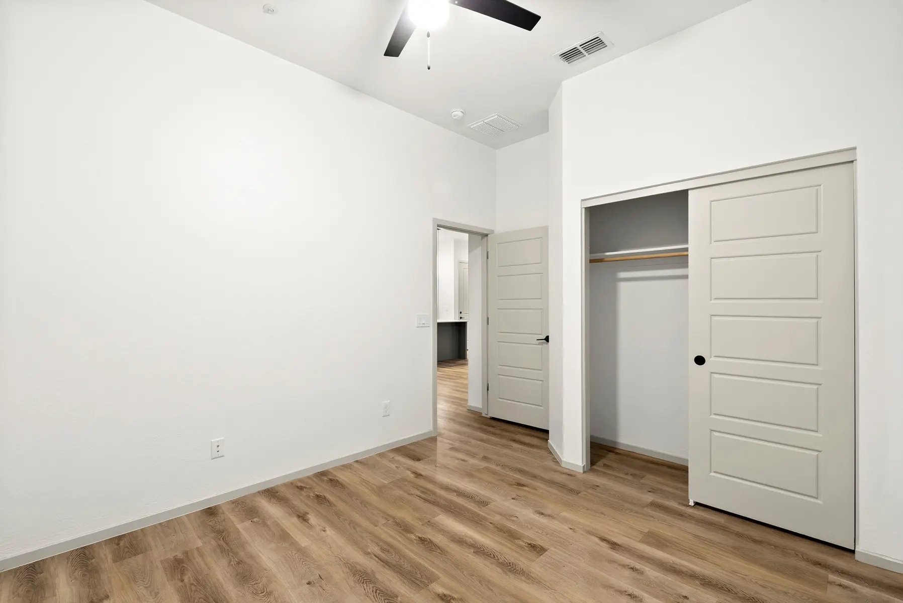 A bright bedroom with light walls, a ceiling fan, and wooden flooring. Two doors leading to a closet and bathroom are visible.