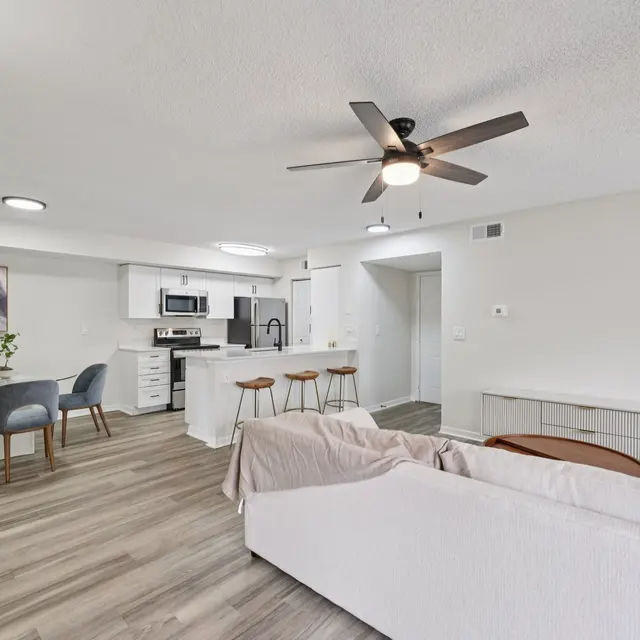 A modern open-concept living space featuring a white sectional sofa, round dining table with four chairs, and a kitchen area with modern appliances. Hardwood flooring throughout, with a ceiling fan above and light-colored walls.