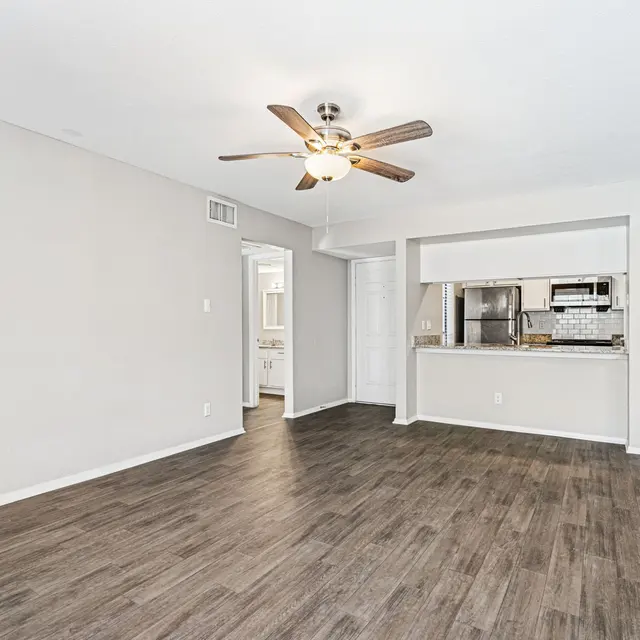 A spacious living room with wood-like flooring and a ceiling fan. The room features light-colored walls, a doorway leading to another room, and a partial view of a kitchen with modern appliances.