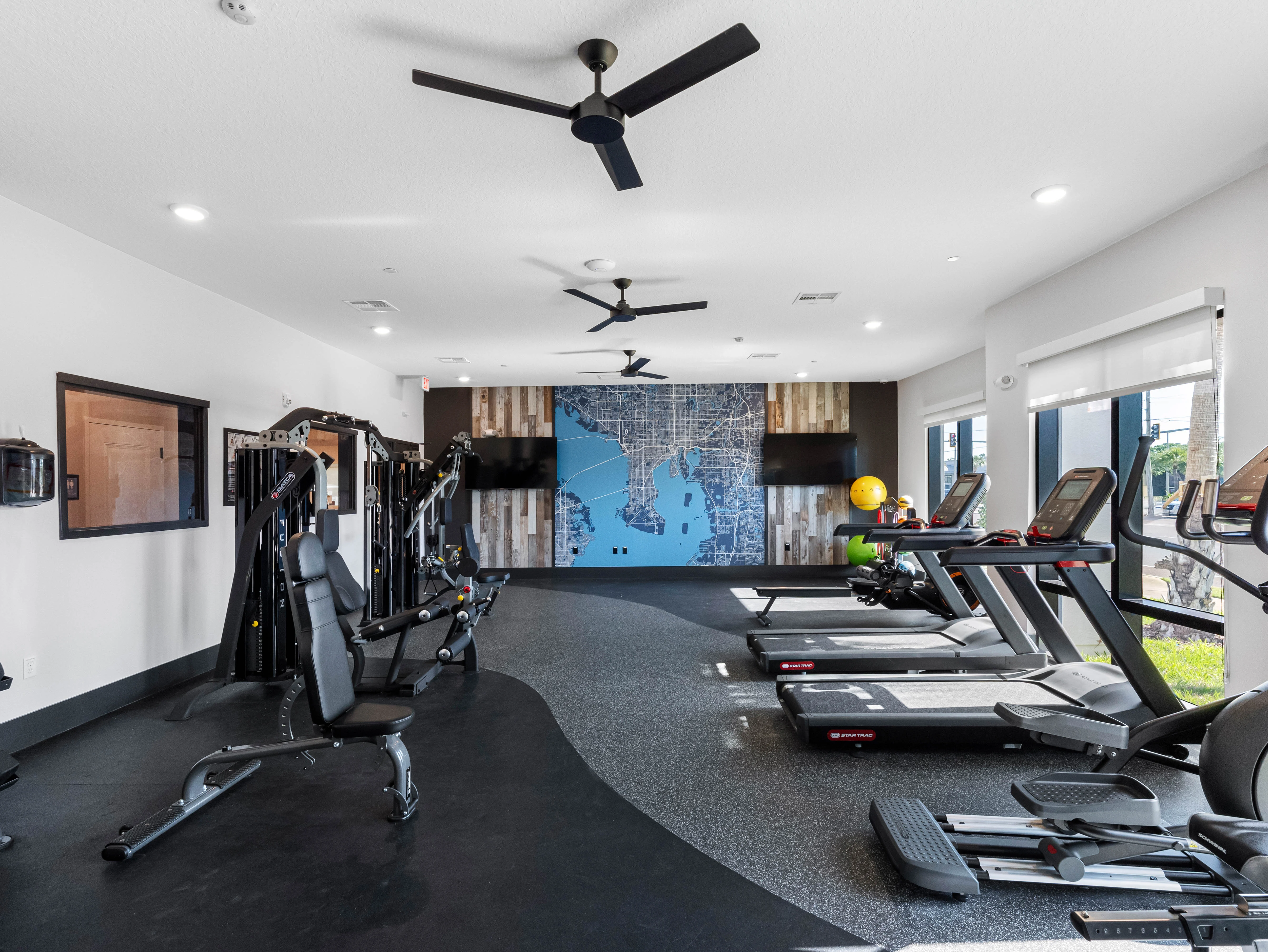 A spacious and well-lit gym featuring exercise machines such as treadmills and weight training equipment. The gym has a stylish mural of a map on one wall and is designed for fitness activities.