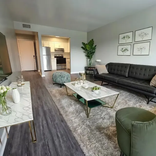 Modern living room featuring a dark sofa, green accents, a marble coffee table, and large TV, with an open kitchen in the background.
