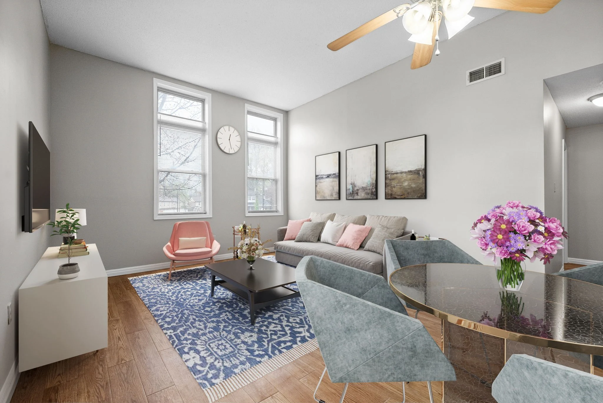 A modern living room featuring a gray sofa with pink cushions, a wooden floor, and a blue patterned rug. There are large windows letting in natural light, a round dining table with gray chairs, and a decorative plant. A coffee table is placed in front of the sofa, and a floral arrangement is on the table.