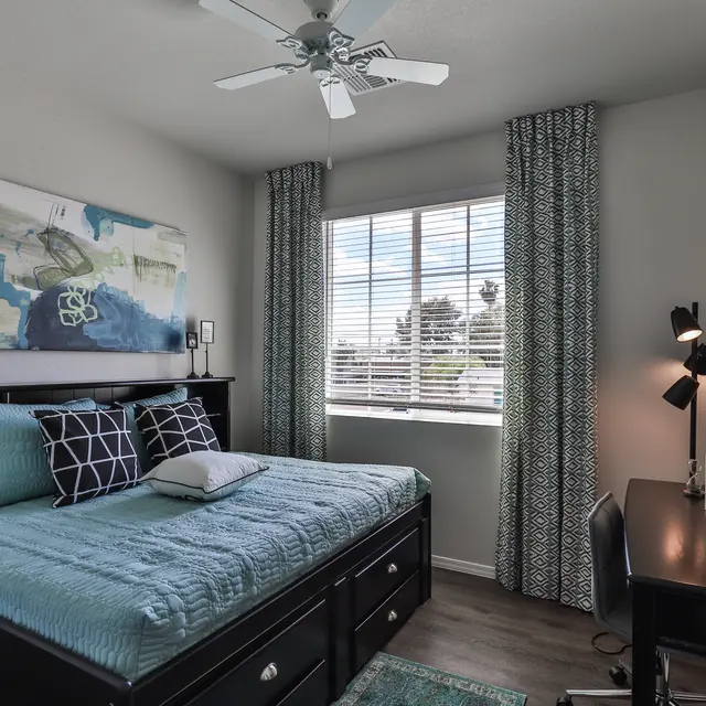 A modern bedroom featuring a bed with a blue bedspread, decorative pillows, and artwork on the walls. The room has a window with light curtains, a ceiling fan, and a black desk with a lamp and decorative items.