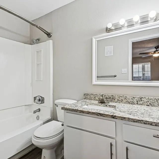 A modern bathroom featuring a white bathtub, a toilet, and a sink with a granite countertop. A large mirror reflects the room's interior, which includes a window and ceiling fan in the background.