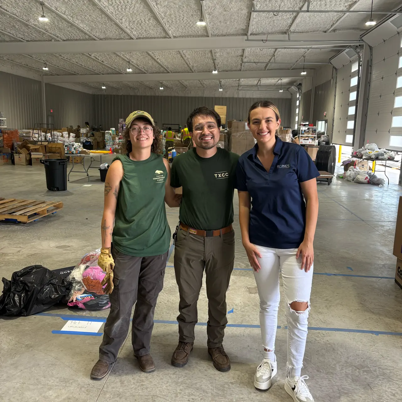 Three people posing together inside a large indoor space filled with boxes and supplies.