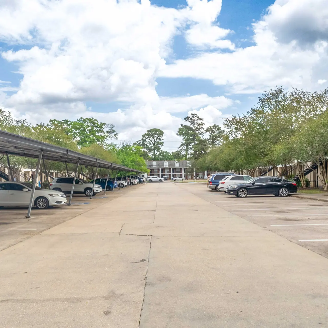An open parking lot with covered parking spaces on either side, flanked by low-rise buildings and greenery. The sky is partly cloudy, with a wide view down the length of the lot.