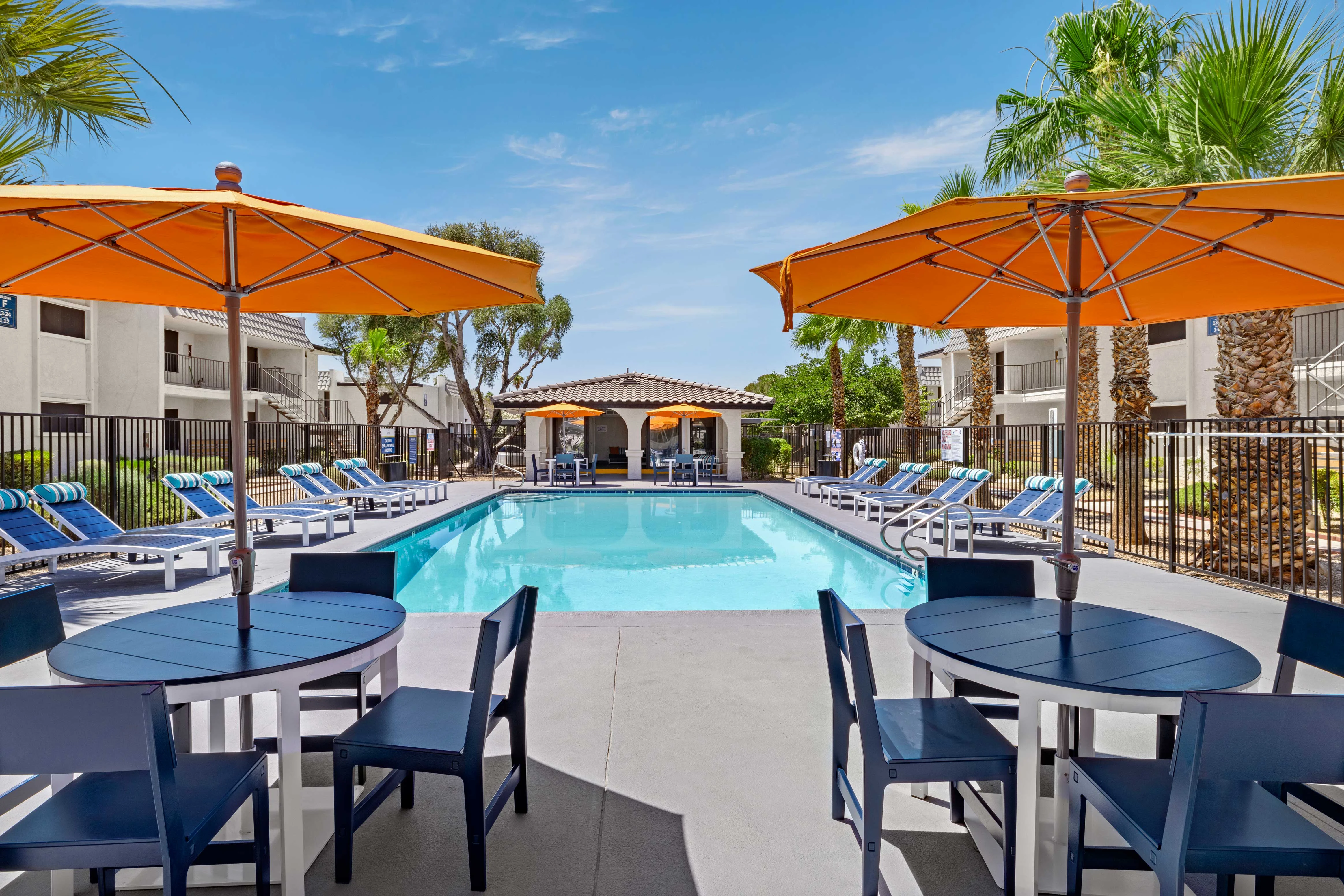A sunny pool area featuring blue lounge chairs and orange umbrellas, with a crystal-clear swimming pool in the background and palm trees lining the space.