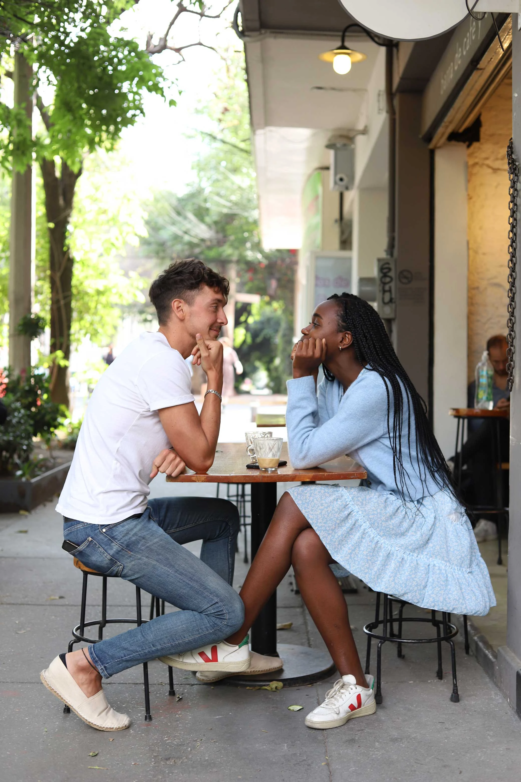 A young couple sits at a small outdoor cafe table, smiling and engaged in conversation, surrounded by greenery.