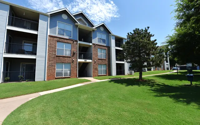 Apartment building with multiple floors, surrounded by green grass and trees, under a clear blue sky.