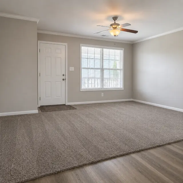 An empty living room featuring light gray walls, a ceiling fan, a window with curtains, and a carpeted floor leading to a wooden section.