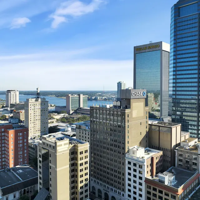 Downtown Jacksonville Skyline A panoramic view of downtown Jacksonville, showcasing a skyline with various buildings, including skyscrapers and mid-rise structures, along with a river in the distance under a clear blue sky.