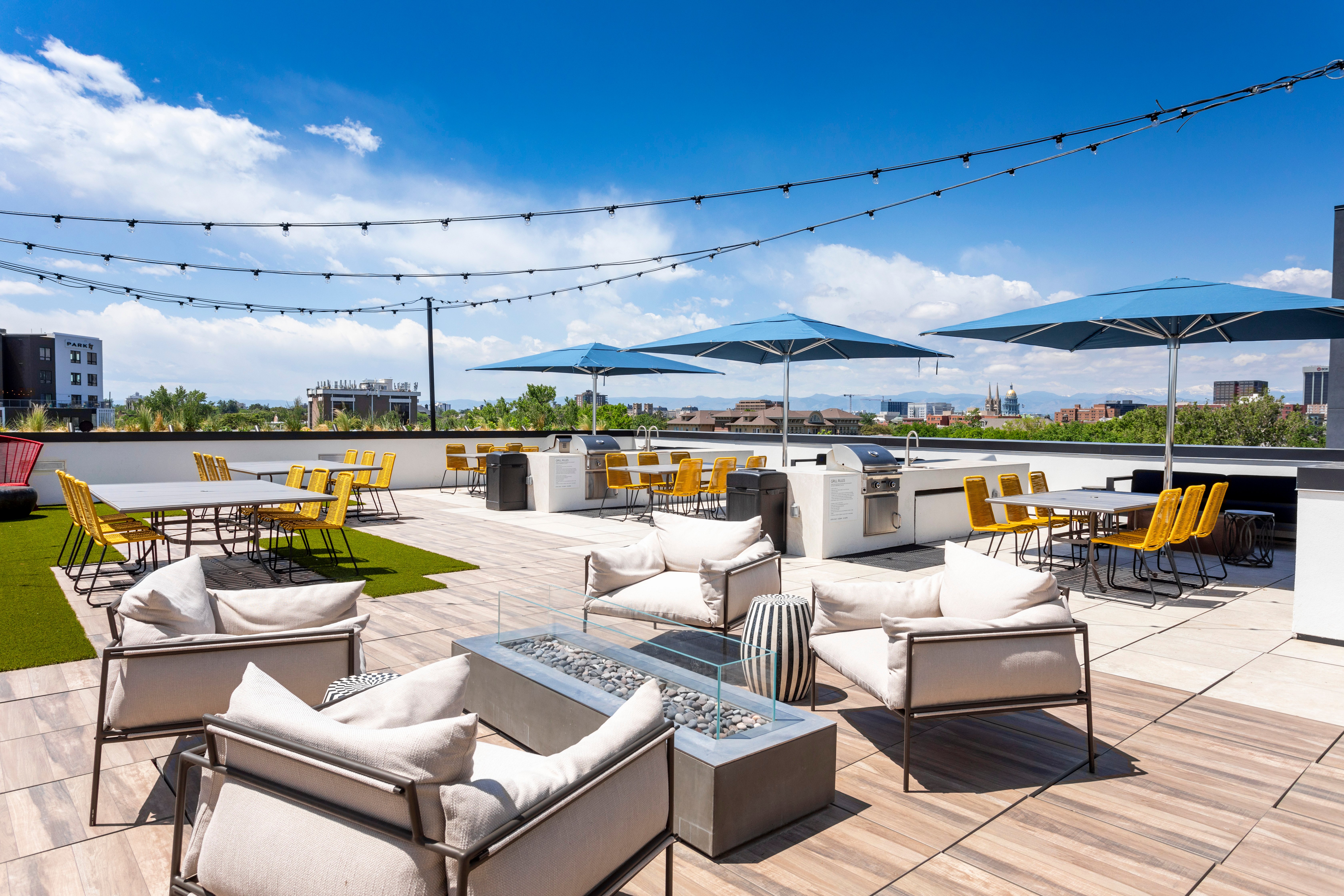 Modern Rooftop Terrace A modern rooftop terrace with seating areas, an outdoor fireplace, and blue umbrellas. The sky is clear and sunny, creating a bright atmosphere.