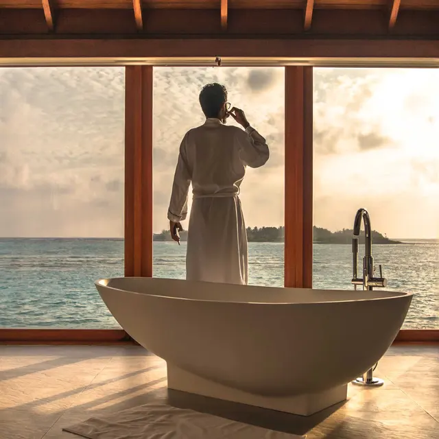 A person in a robe stands by a large window overlooking the ocean, with a freestanding bathtub nearby. Natural light floods the room.