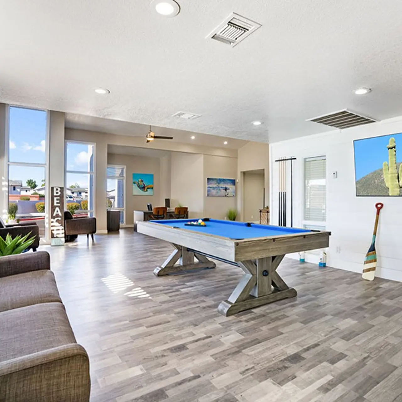 A spacious game room featuring a blue pool table, a cozy gray couch, and large windows allowing natural light to flood in. Decor includes a wall-mounted artwork of a cactus and beach-themed items.