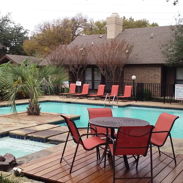 A swimming pool area with lounge chairs and a table set near the pool, surrounded by greenery and a small rock feature.