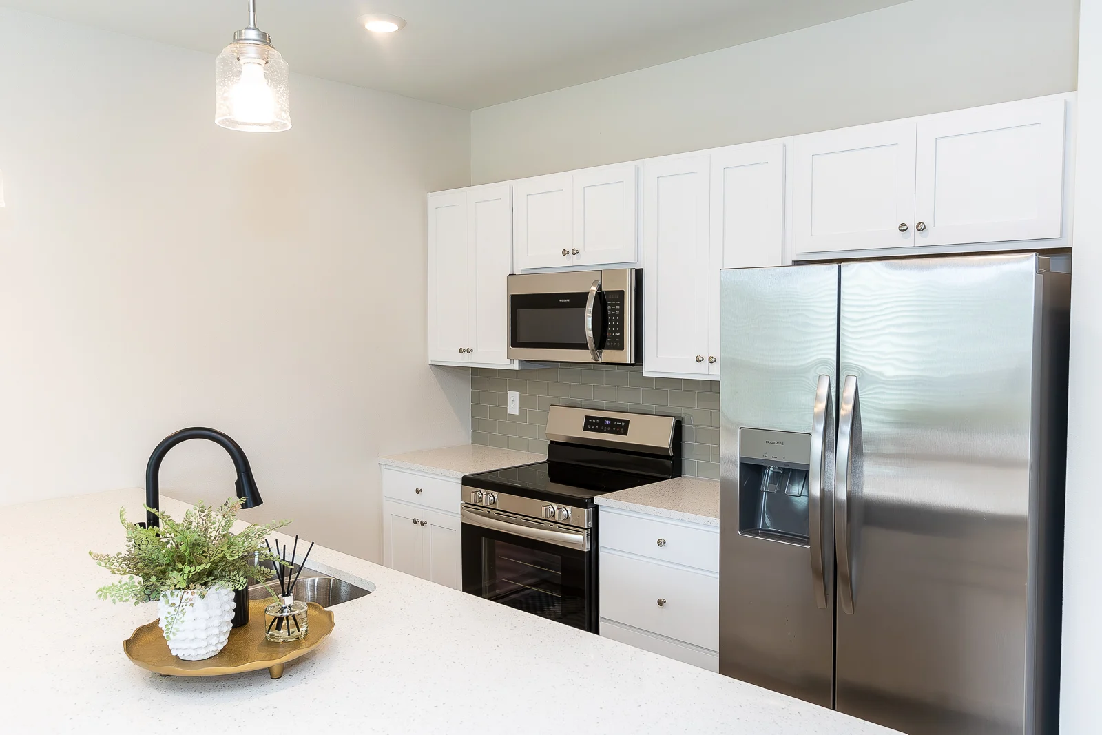 A modern kitchen featuring white cabinetry, stainless steel appliances, and elegant decor. The kitchen has a large island with a sink and decorative plant, and overhead lighting adds warmth to the space.