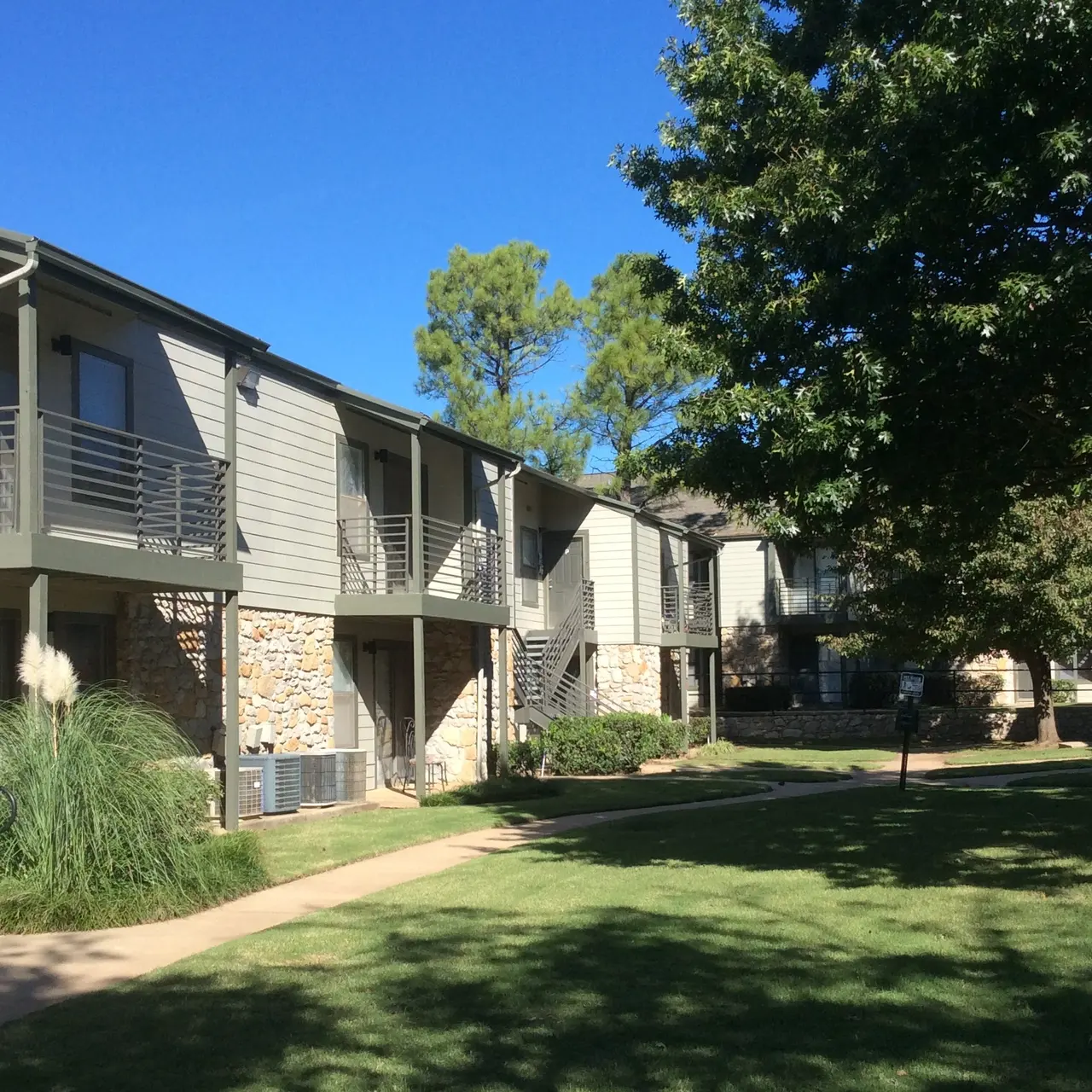 The Hunt A two-story apartment complex with stone and siding exterior, balconies, surrounded by greenery and trees on a sunny day.