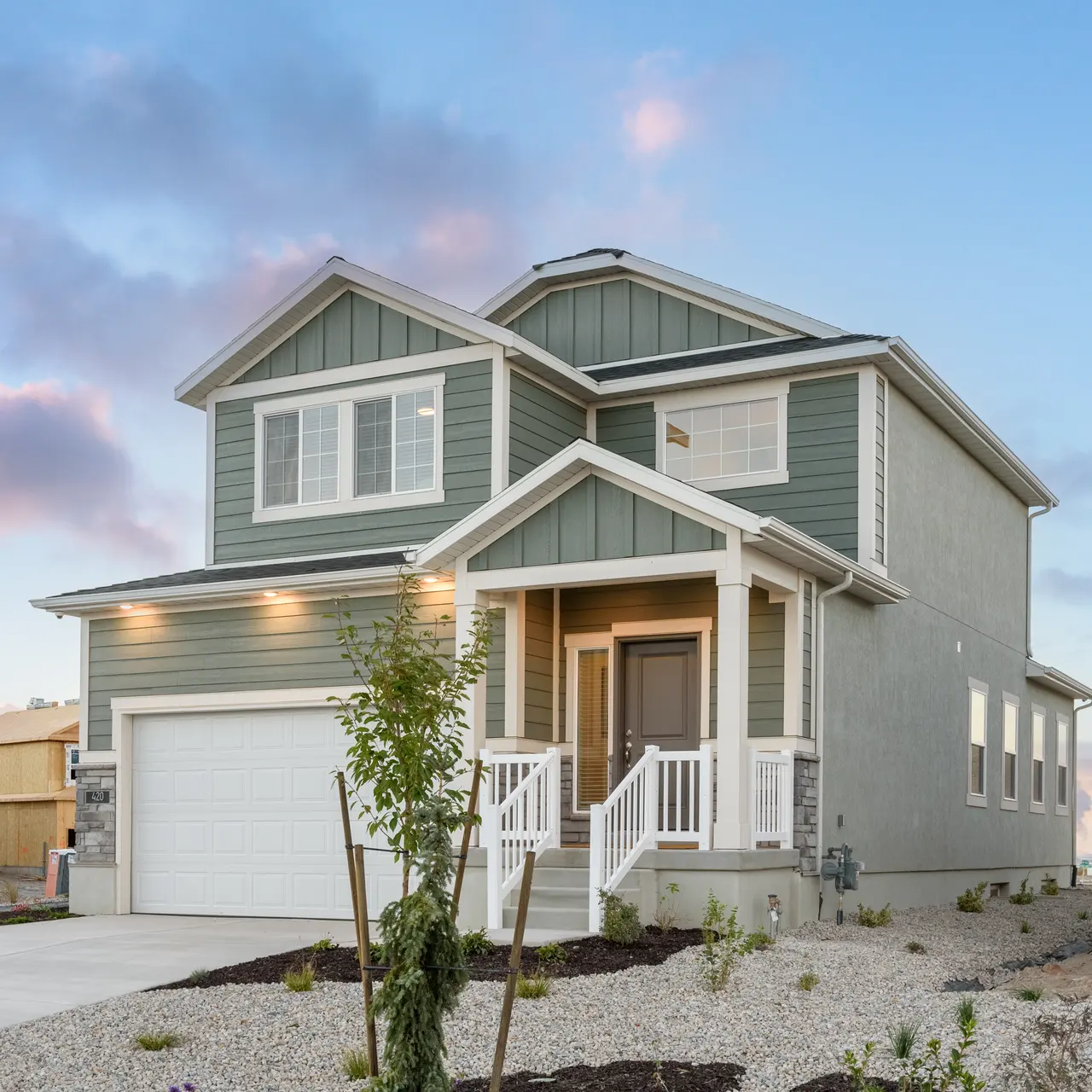 A modern two-story house with a light green exterior and white accents, featuring a garage and a small landscaped front yard.